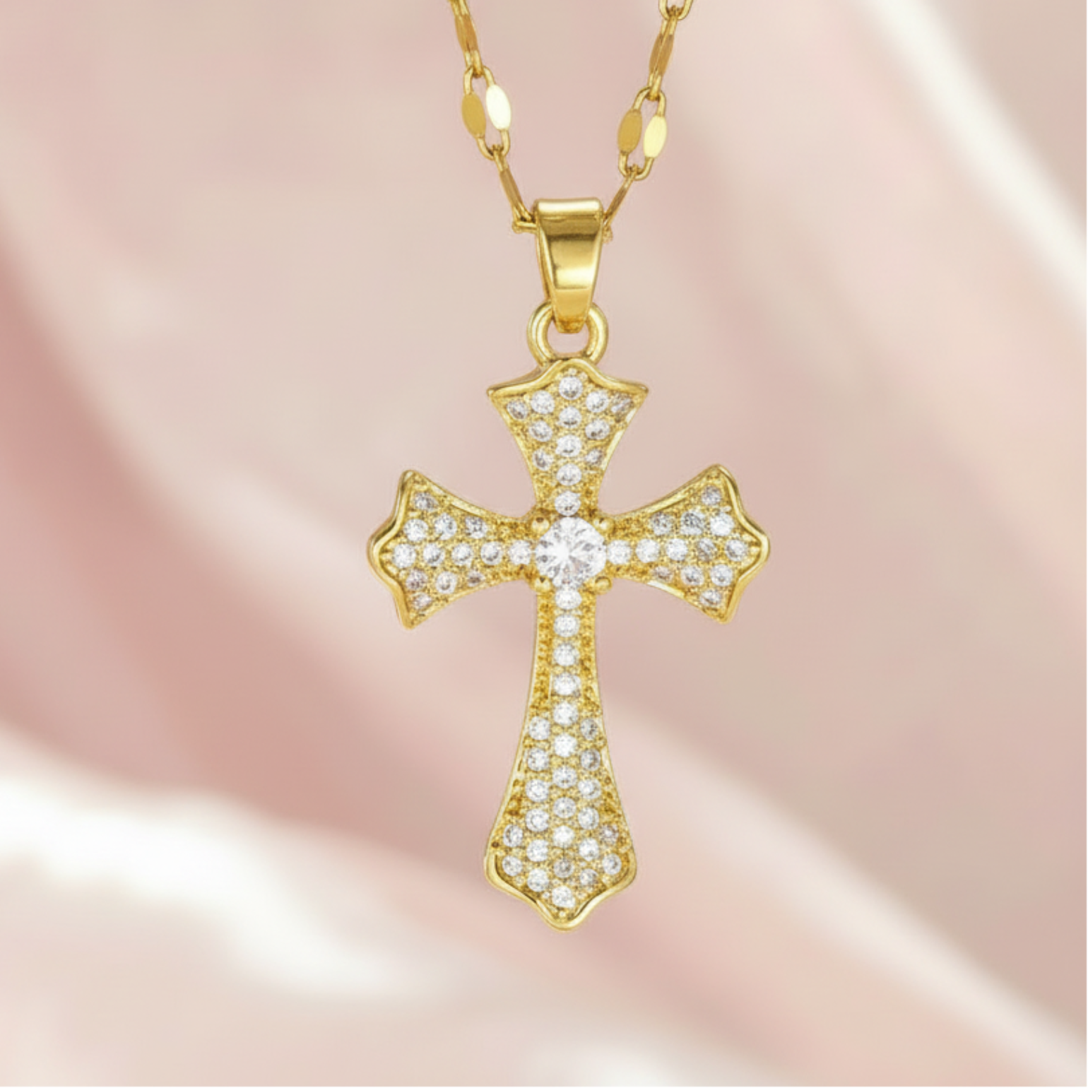 A gold cross pendant necklace paved with small clear stones, hanging from a gold chain against a soft pink satin background.