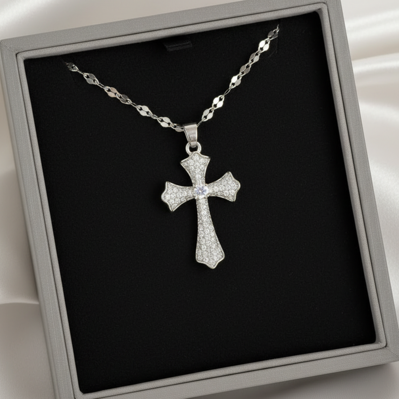 A silver cross pendant necklace with a pavé-style textured finish, displayed inside a gray presentation box with a black insert on a white satin background.