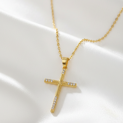 A gold cross pendant necklace with small clear stones set along the horizontal arm and lower vertical arm, displayed on draped white satin fabric.
