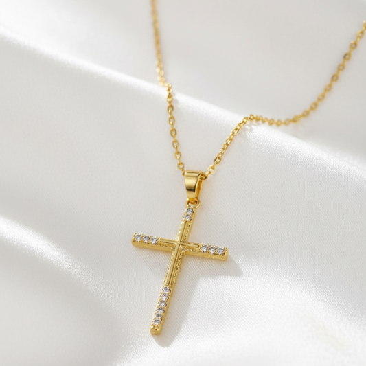 A gold cross pendant necklace with small clear stones set along the horizontal arm and lower vertical arm, displayed on draped white satin fabric.