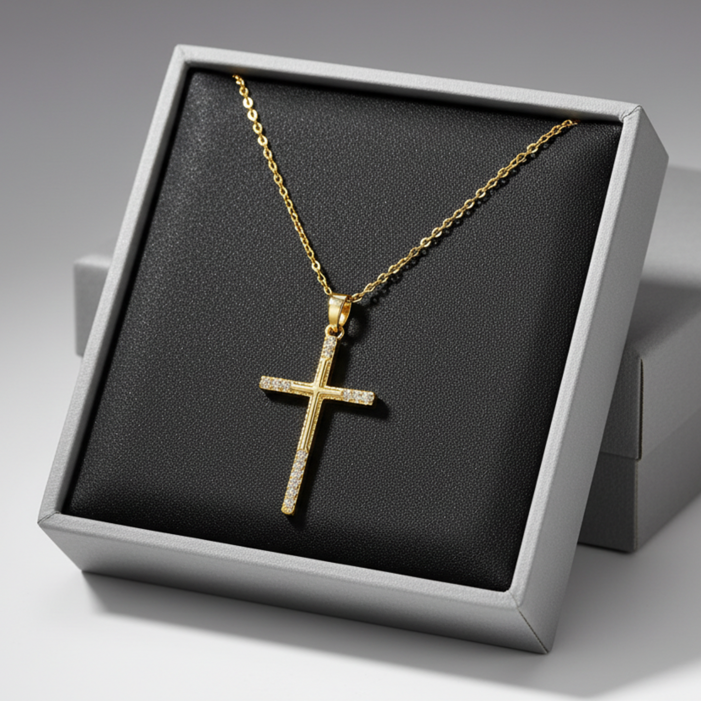 A gold cross pendant necklace with small clear stones set along the arms, displayed on a grey jewelry box.
