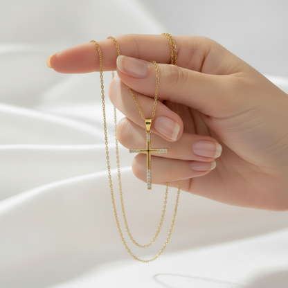 A hand with manicured nails holds a delicate gold cross pendant necklace adorned with small clear stones, set against a soft white draped fabric background.