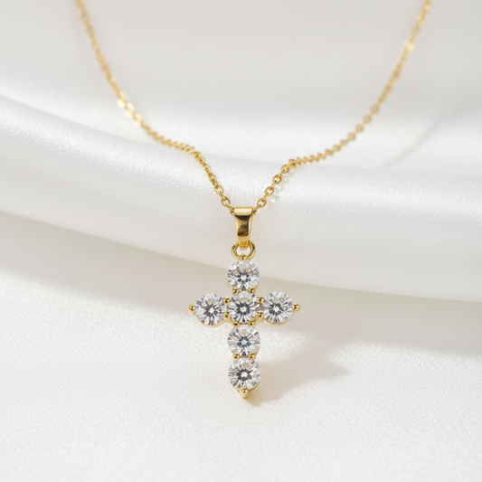 A gold cross pendant necklace adorned with clear gemstones, displayed on a white satin fabric background.
