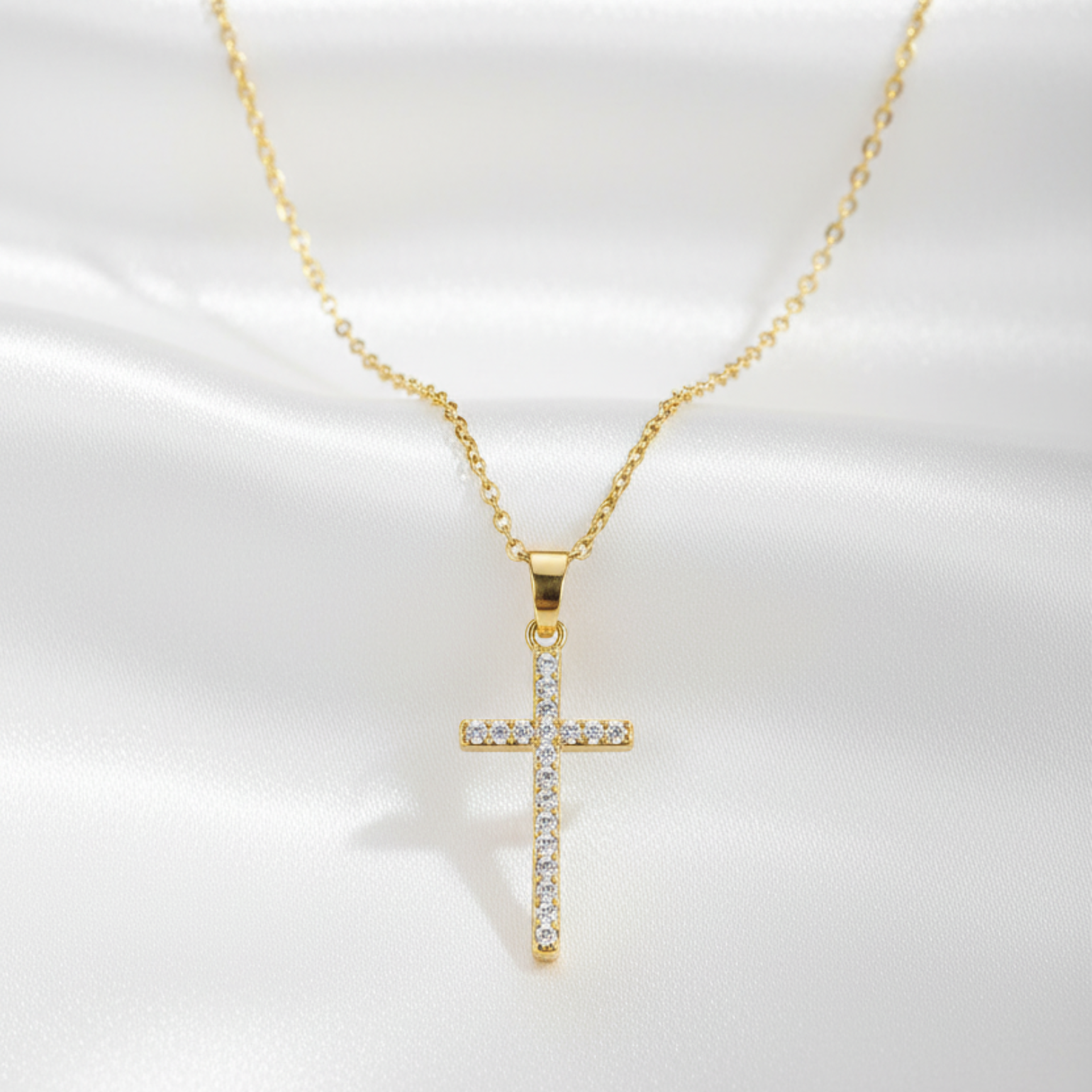 A gold cross pendant necklace adorned with small clear stones, displayed on a white satin fabric background.