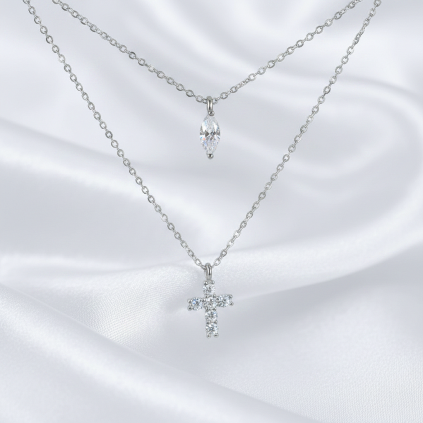 Two delicate silver-tone necklaces displayed on a white satin fabric background. The top necklace features a small silver chain with a marquise-cut clear crystal pendant, while the bottom necklace has a longer chain with a small silver cross pendant set with clear crystals.