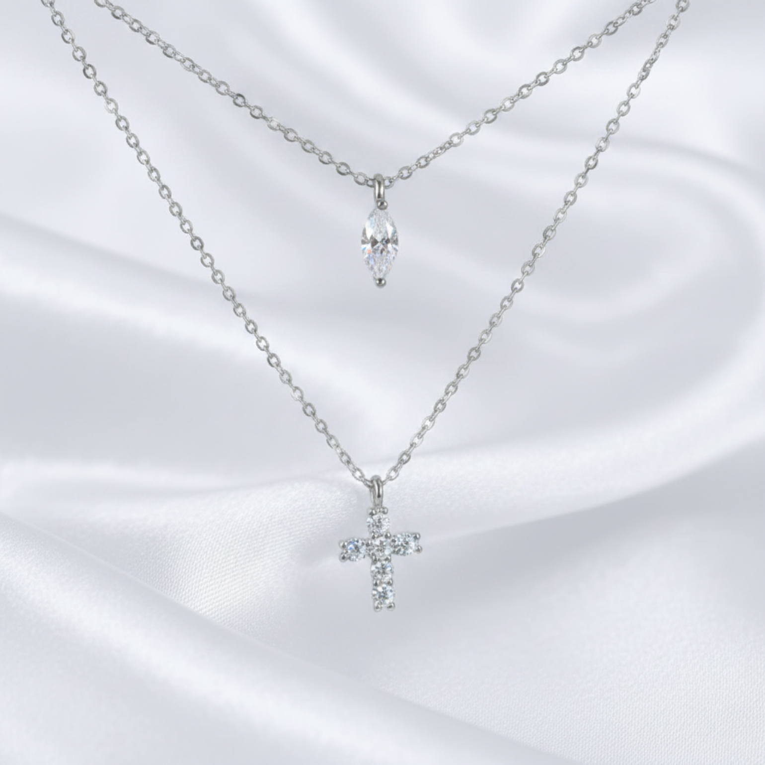 Two delicate silver-tone necklaces displayed on a white satin fabric background. The top necklace features a small silver chain with a marquise-cut clear crystal pendant, while the bottom necklace has a longer chain with a small silver cross pendant set with clear crystals.