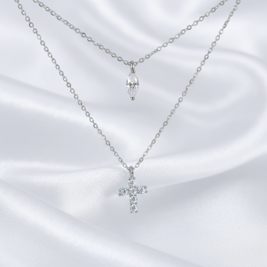 Two delicate silver-tone necklaces displayed on a white satin fabric background. The top necklace features a small silver chain with a marquise-cut clear crystal pendant, while the bottom necklace has a longer chain with a small silver cross pendant set with clear crystals.